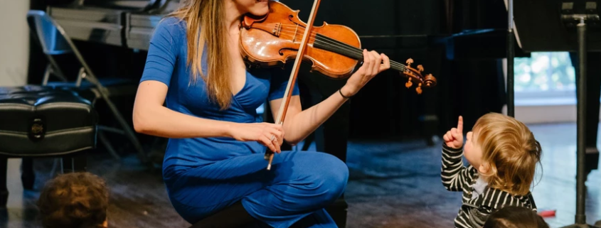 A woman kneels while playing violin for onlooking children
